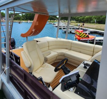 Interior view of a Canalake Double Decker boat featuring beige seating, steering wheel, and a scenic view of the water and docked boats.